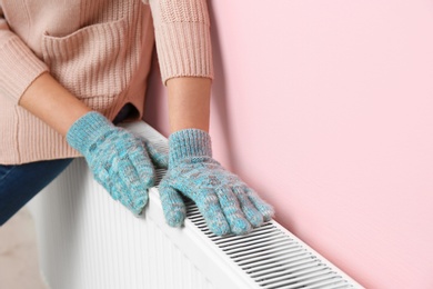 Photo of Woman in gloves warming hands on heating radiator near color wall. Space for text