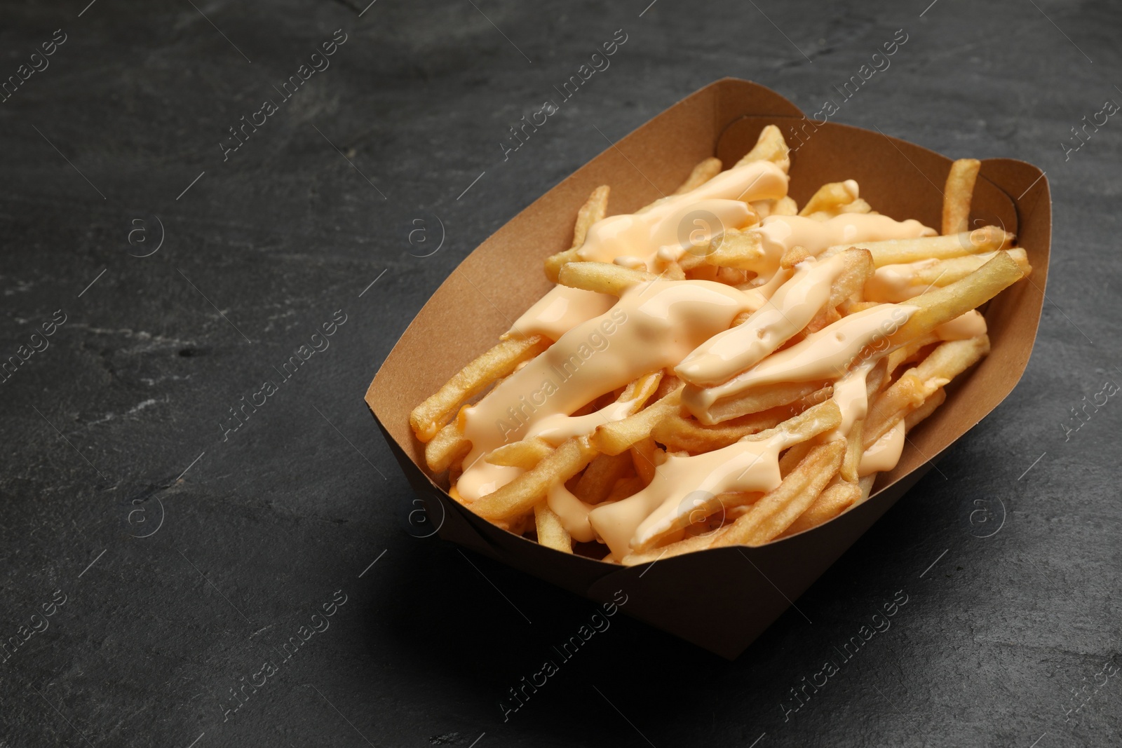 Photo of Tasty potato fries and cheese sauce in paper container on black table, closeup