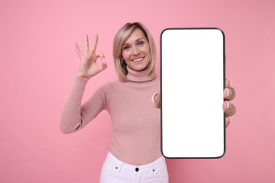 Happy woman showing mobile phone with blank screen on pink background. Mockup for design