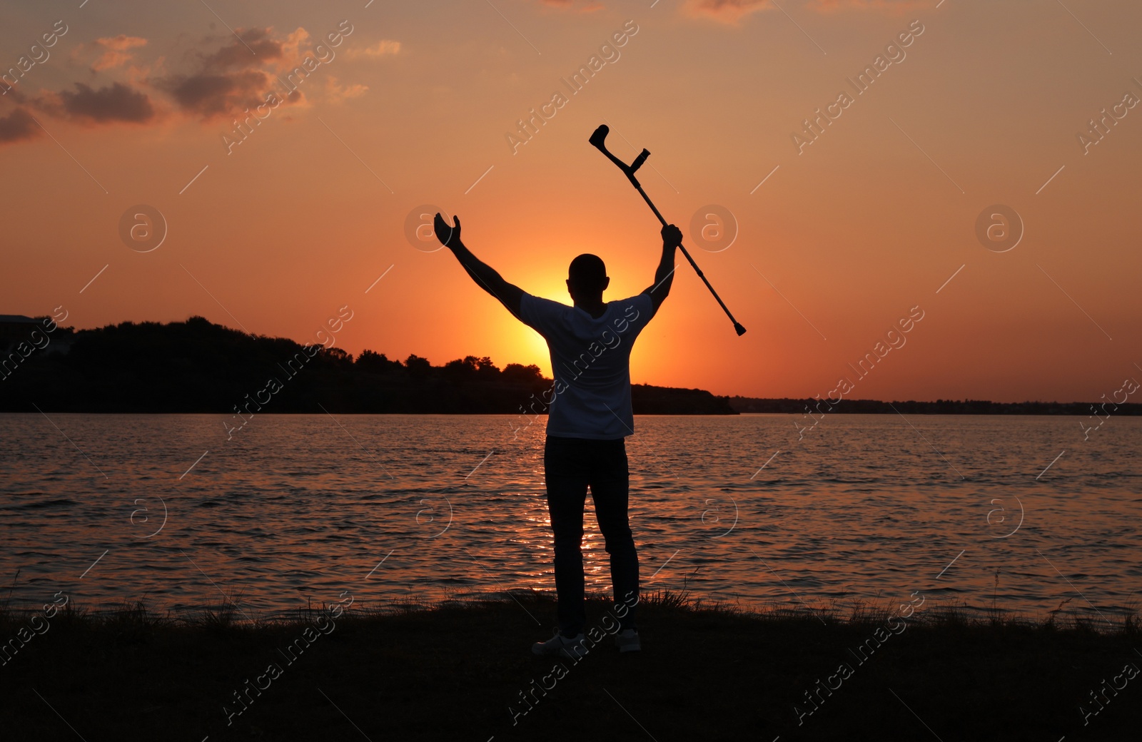 Photo of Man raising elbow crutch up to sky near river at sunset, back view. Healing miracle