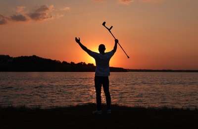 Photo of Man raising elbow crutch up to sky near river at sunset, back view. Healing miracle