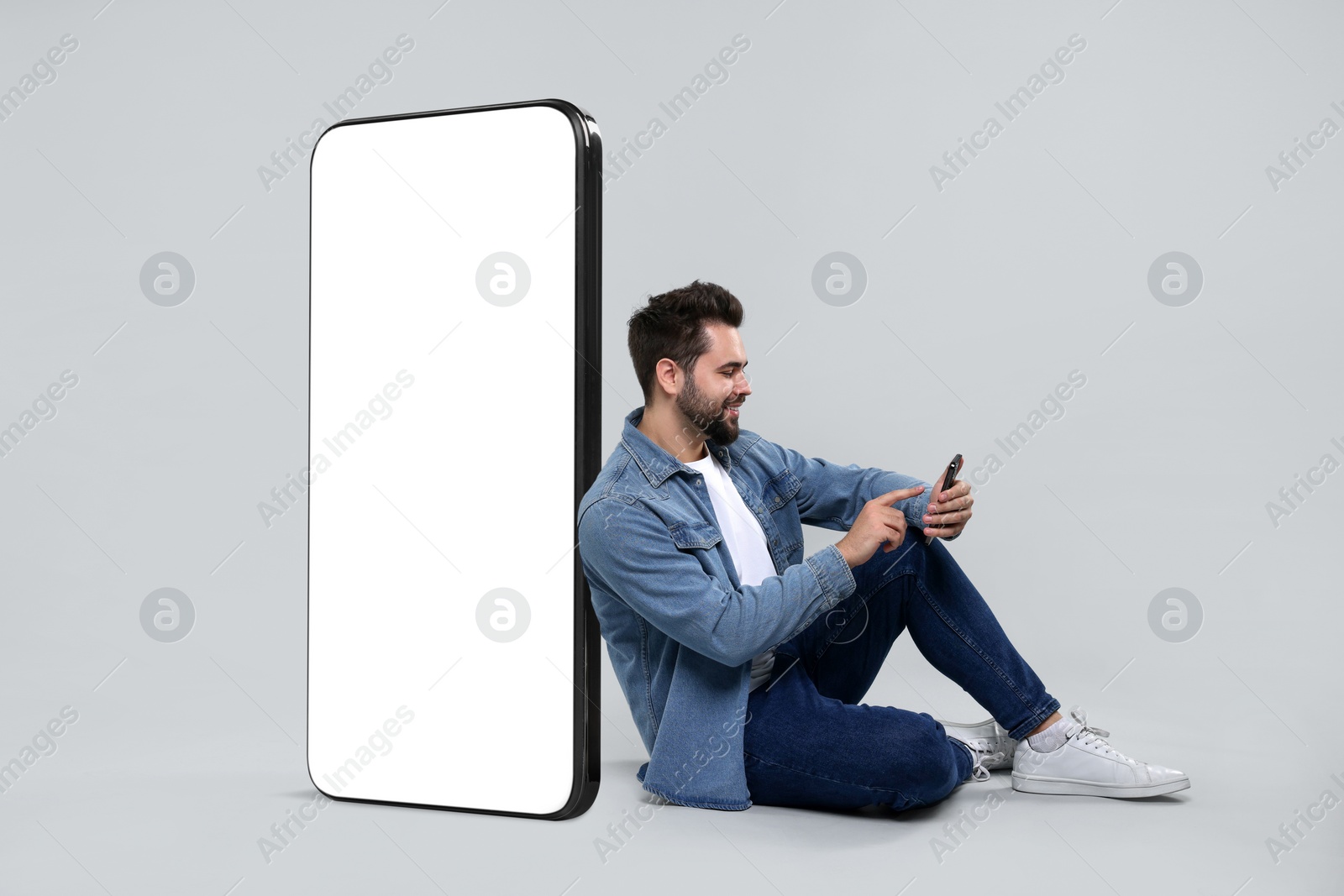 Image of Man with mobile phone sitting near huge device with empty screen on grey background. Mockup for design