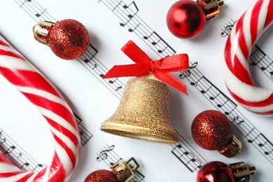 Photo of Golden shiny bell with red bow, candy canes and decorative Christmas baubles on music sheet, flat lay