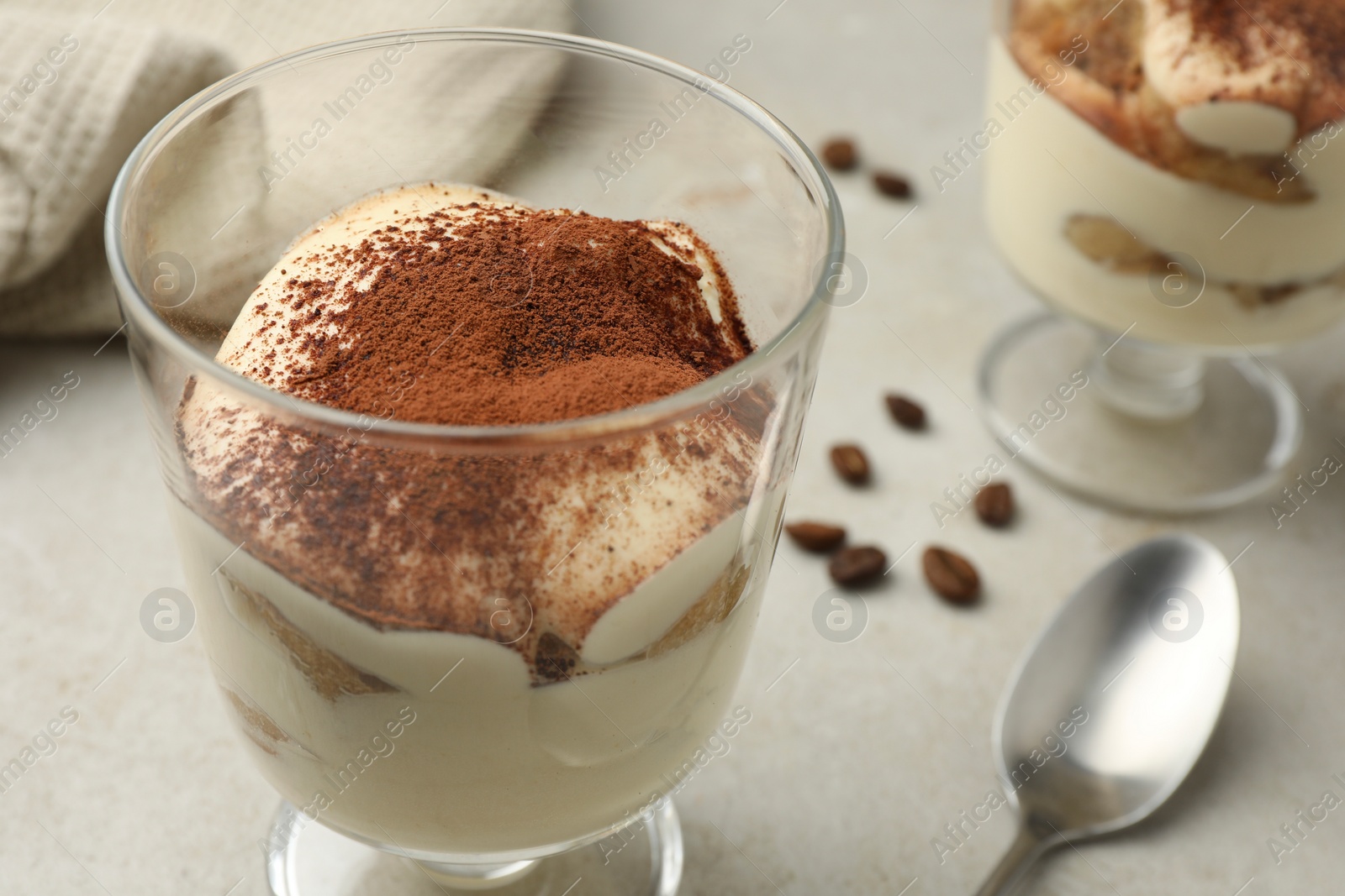 Photo of Delicious tiramisu in glass, spoon and scattered coffee beans on light table, closeup