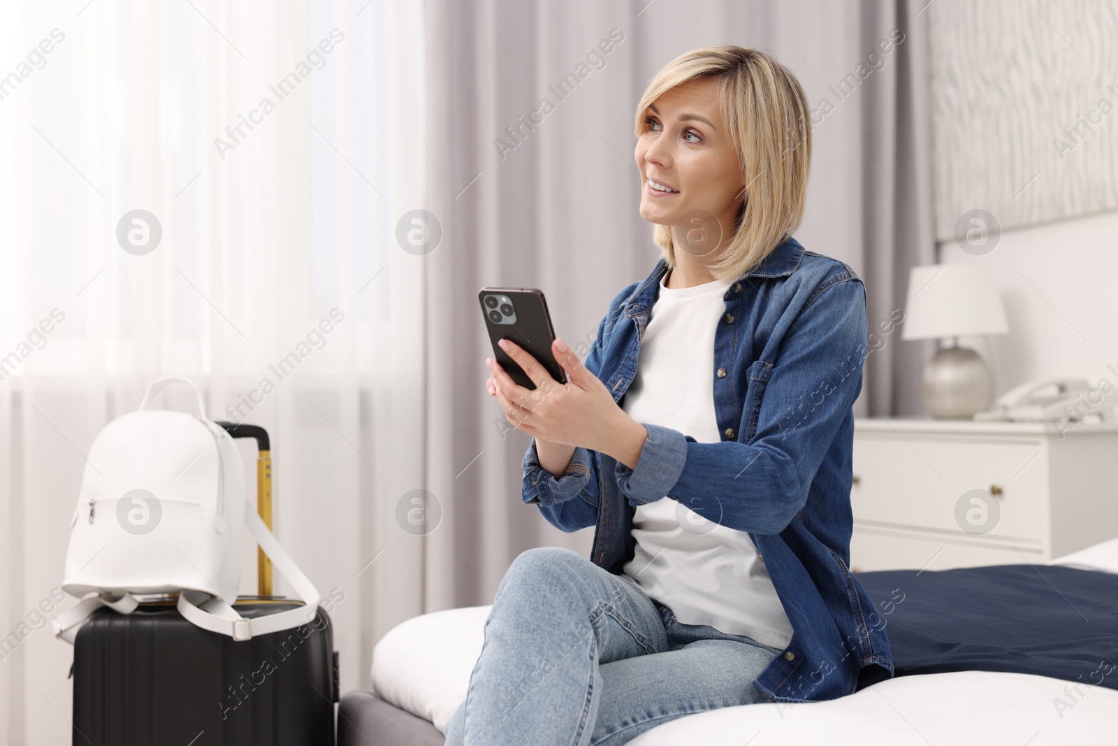 Photo of Smiling guest with smartphone relaxing on bed in stylish hotel room