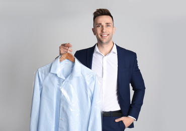 Man holding hanger with shirt in plastic bag on light grey background. Dry-cleaning service