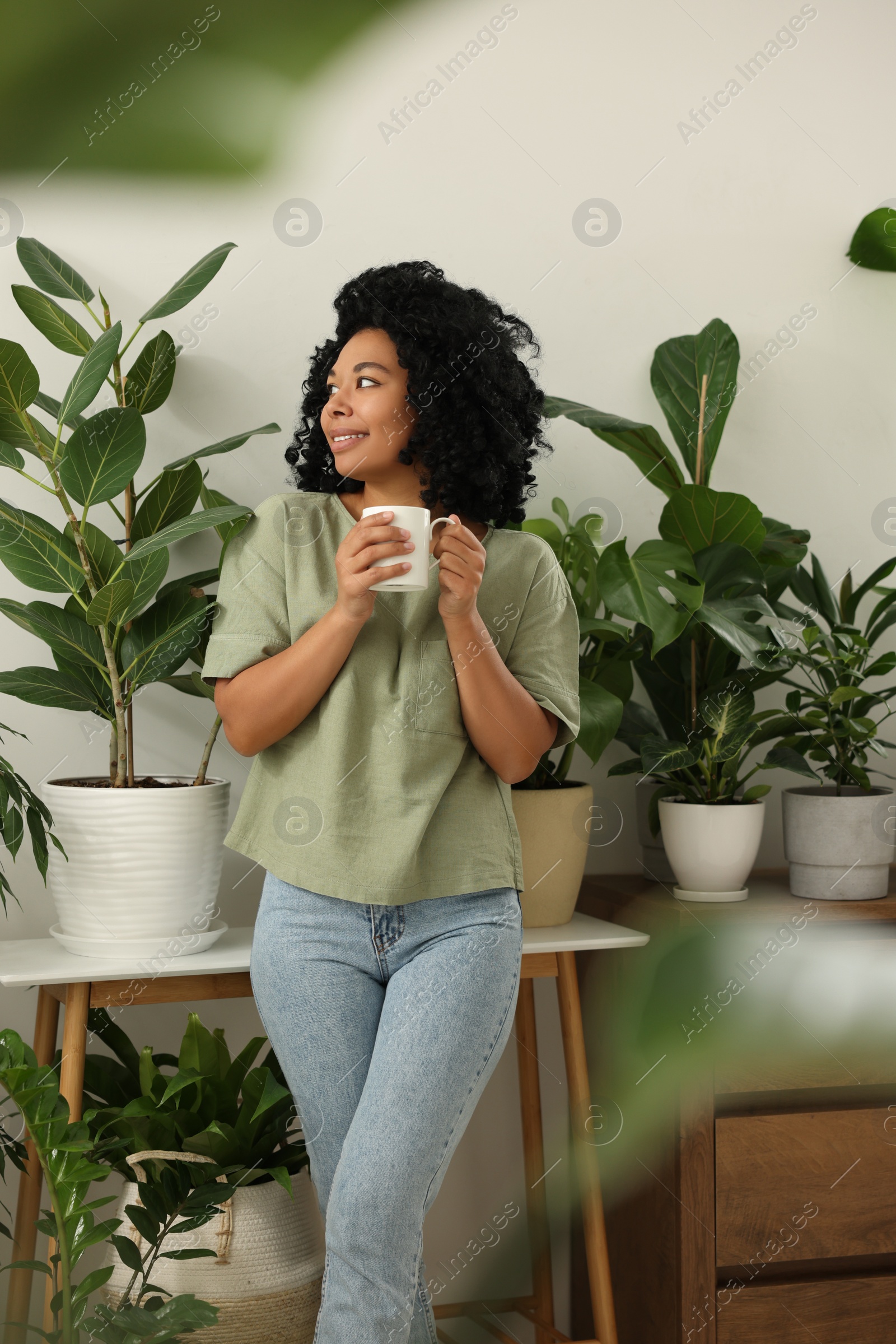 Photo of Relaxing atmosphere. Happy woman with cup of hot drink near beautiful houseplants in room