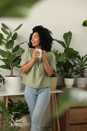 Photo of Relaxing atmosphere. Happy woman with cup of hot drink near beautiful houseplants in room