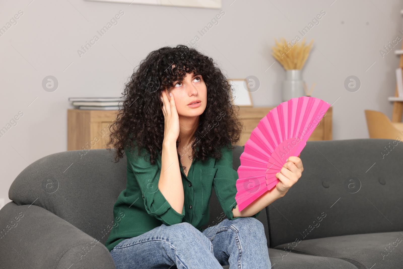 Photo of Young woman waving pink hand fan to cool herself on sofa at home