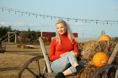 Beautiful woman sitting on wooden cart with pumpkins and hay in field. Autumn season