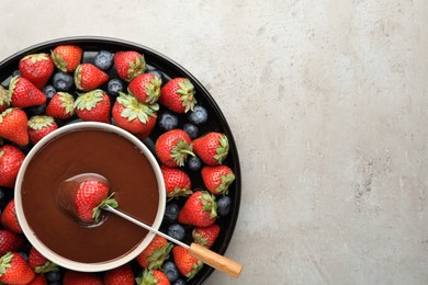 Fondue fork with strawberry in bowl of melted chocolate surrounded by different berries on light table, top view. Space for text