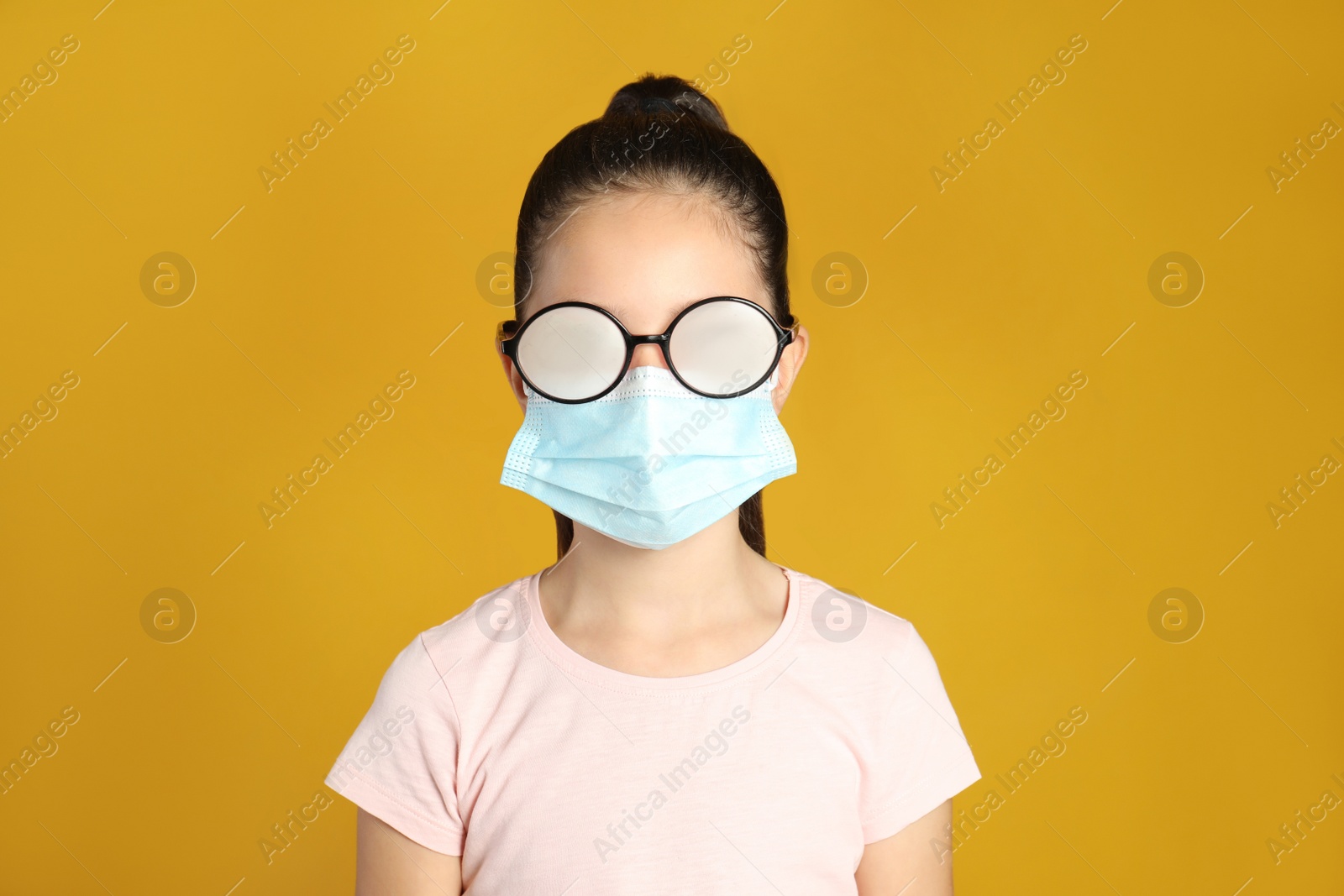 Photo of Little girl with foggy glasses caused by wearing medical face mask on yellow background. Protective measure during coronavirus pandemic