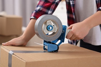 Photo of Woman applying adhesive tape on box with dispenser indoors, closeup