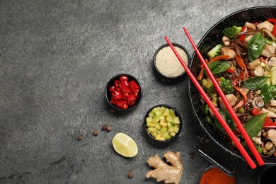 Photo of Stir-fry. Tasty noodles with meat in wok, chopsticks and ingredients on grey textured table, flat lay. Space for text