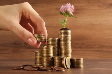 Photo of Woman putting coin onto stack with green plant at wooden table, closeup. Investment concept