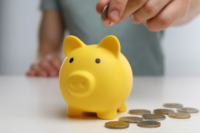 Man putting coin into yellow piggy bank at white table, closeup