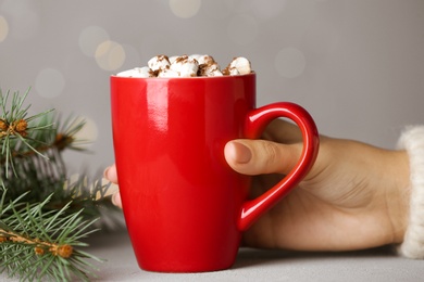 Photo of Woman holding cup of tasty cocoa with marshmallows at white table against blurred lights, closeup