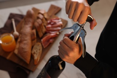 Photo of Romantic dinner. Man opening wine bottle with corkscrew at table indoors, closeup