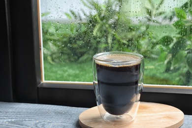 Photo of Glass of coffee on wooden windowsill, space for text. Rainy weather