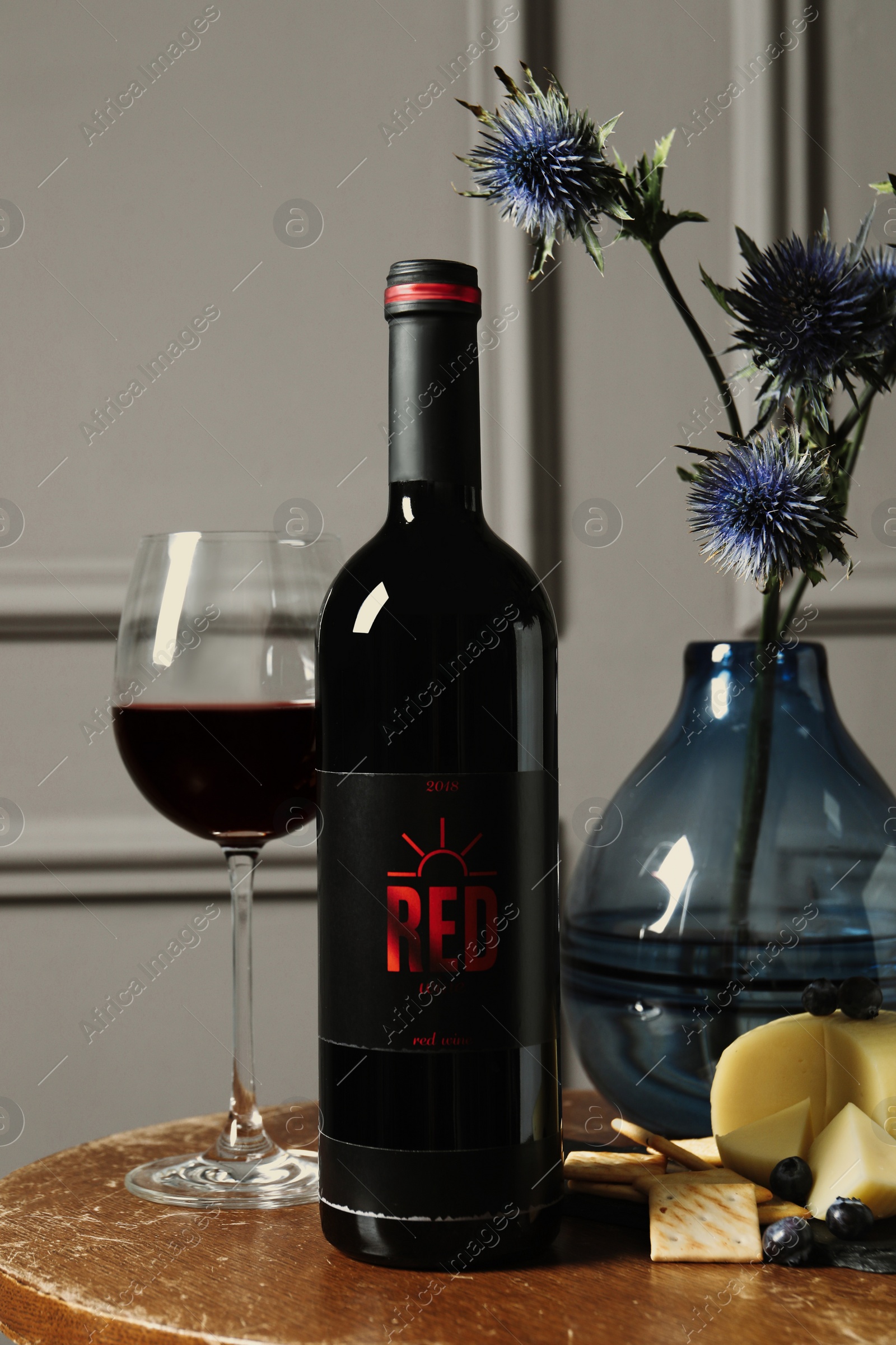 Photo of Bottle of red wine, glass, delicious snacks and beautiful bouquet on wooden table indoors