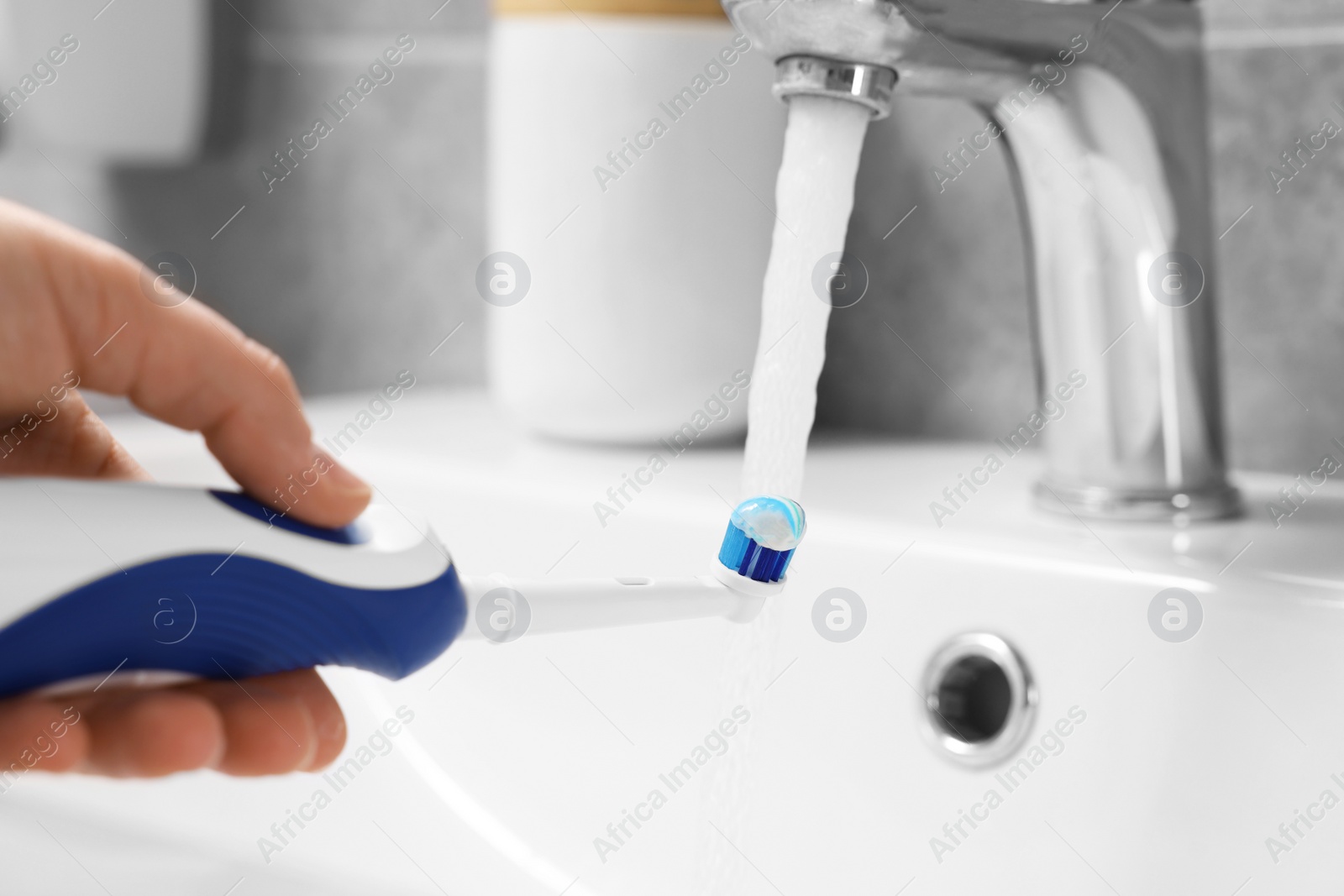 Photo of Woman holding electric toothbrush with paste near flowing water above sink in bathroom, closeup