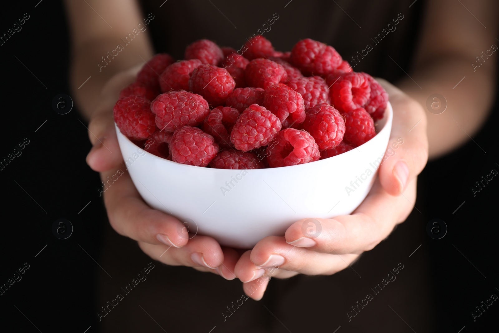 Photo of Woman holding bowl of delicious ripe raspberries on black background, closeup