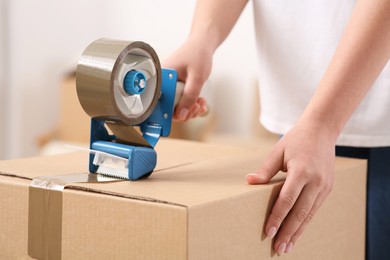 Photo of Woman applying adhesive tape on box with dispenser indoors, closeup