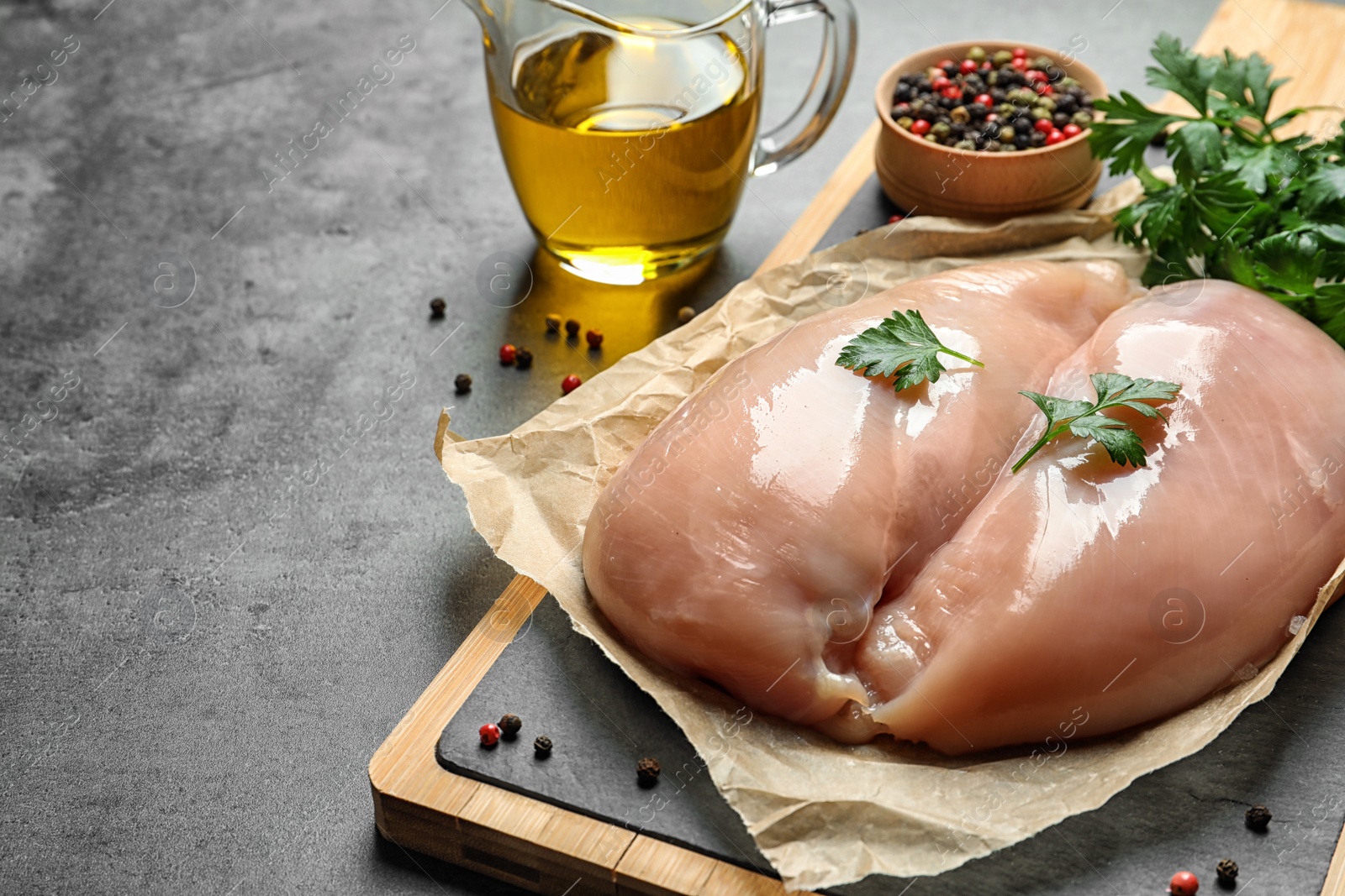 Photo of Wooden board with raw chicken breasts, pepper and parsley near olive oil in pitcher on table