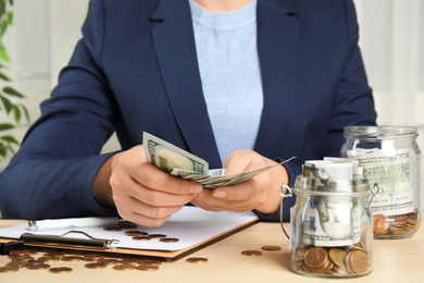 Woman counting money near glass jar with dollar bills and coins on wooden table, closeup