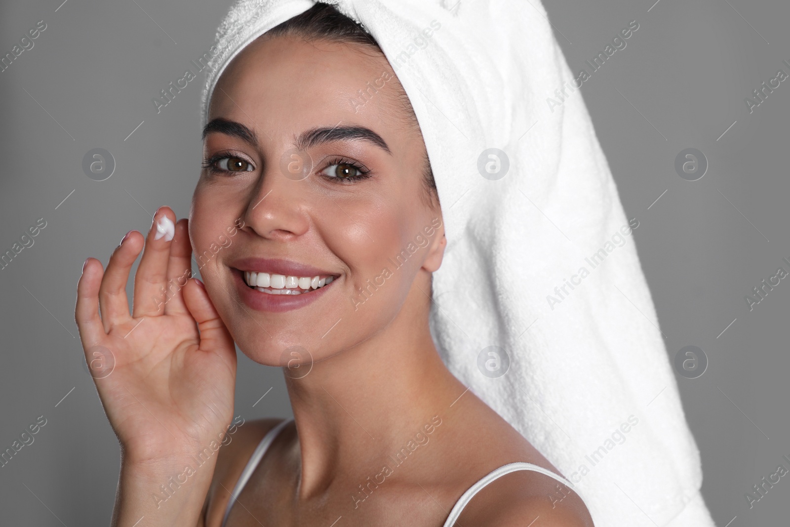 Photo of Beautiful young woman with towel applying cream on face against grey background