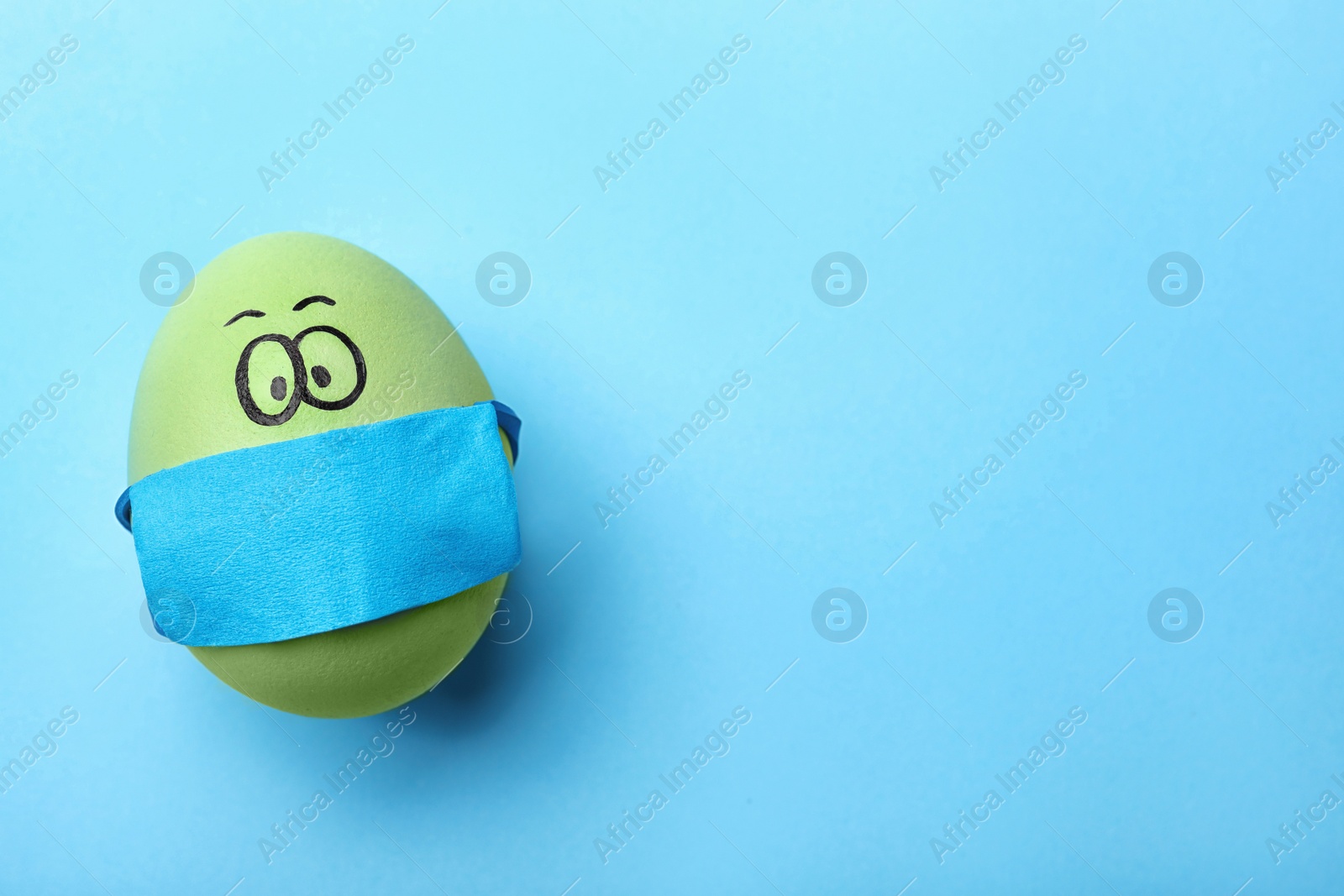 Photo of Bright egg in protective mask on light blue background, top view and space for text. Easter holiday during coronavirus quarantine