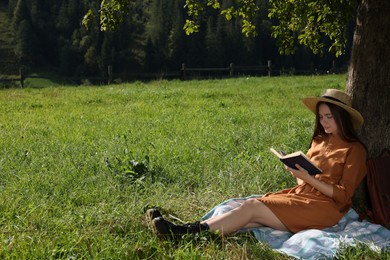 Young woman reading book under tree on meadow near forest