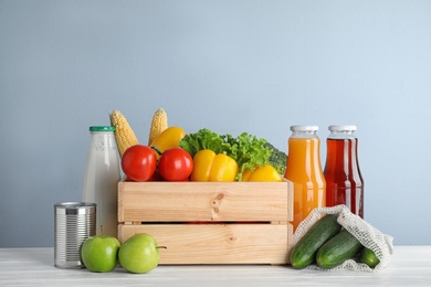 Crate with fresh vegetables and other products on white wooden table against light blue background
