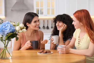 Photo of Happy young friends with cups of drink spending time together at table in kitchen