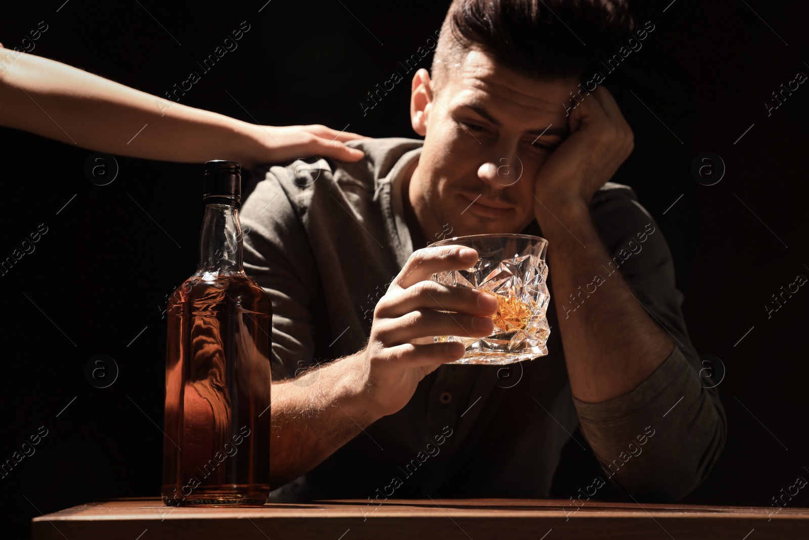 Photo of Addicted man at wooden table against black background, focus on glass of alcoholic drink