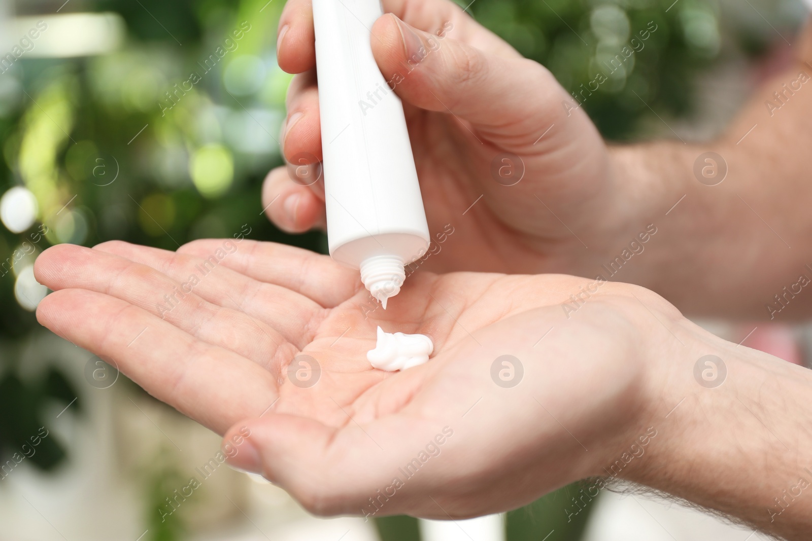 Photo of Man applying moisturizing cream from tube onto hand on blurred background, closeup