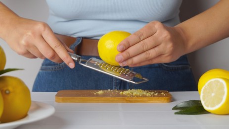 Photo of Woman grating lemon zest on wooden board at white tiled table, closeup