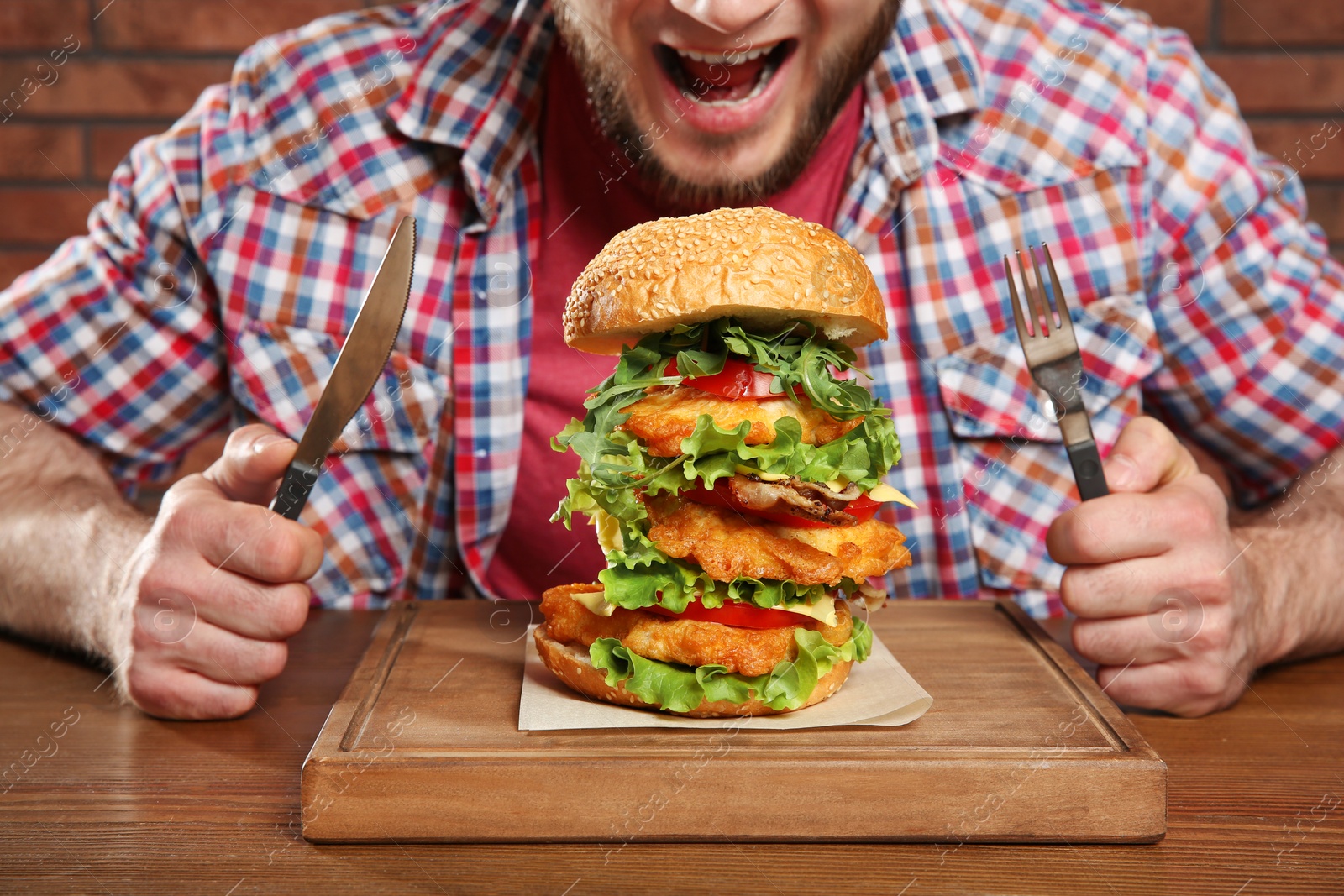 Photo of Young hungry man with cutlery and tasty huge burger at table