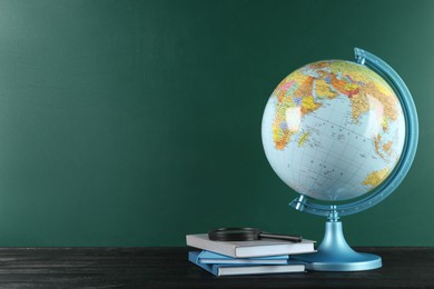 Globe, books and magnifying glass on black wooden table near green chalkboard, space for text. Geography lesson