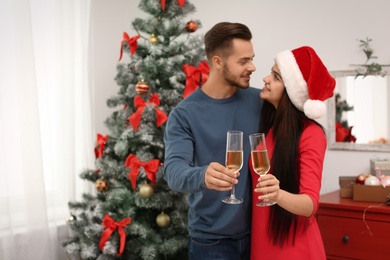 Photo of Young beautiful couple with glasses of champagne near Christmas tree at home