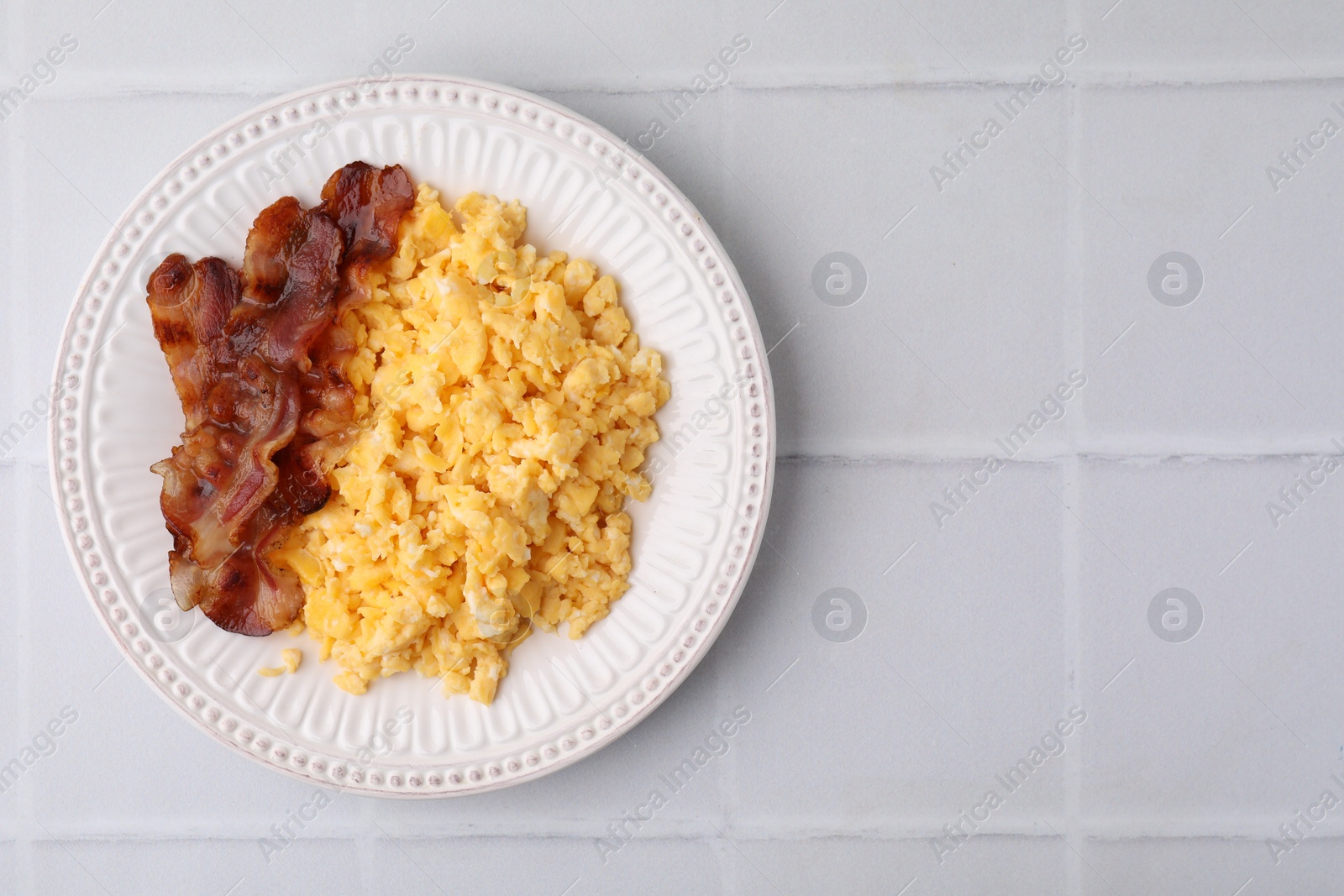 Photo of Delicious scrambled eggs with bacon in plate on white tiled table, top view. Space for text