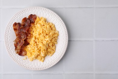 Delicious scrambled eggs with bacon in plate on white tiled table, top view. Space for text