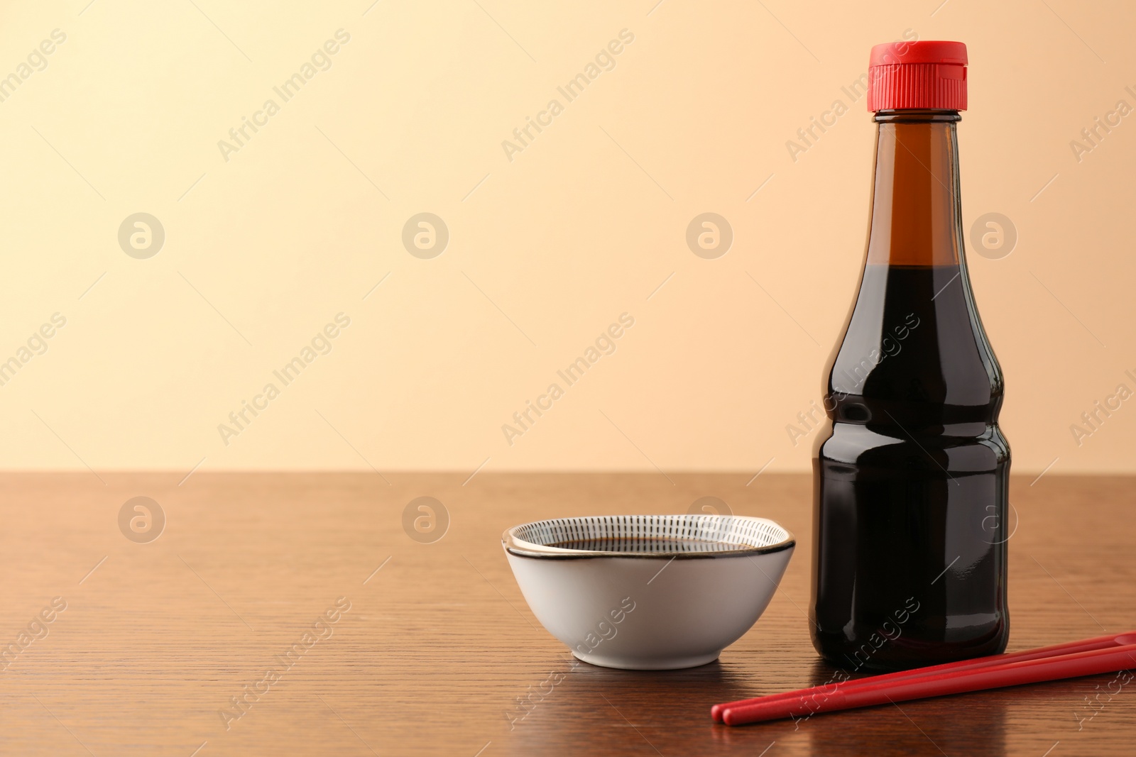 Photo of Bottle, bowl with soy sauce and chopsticks on wooden table against beige background. Space for text