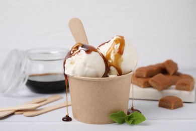 Photo of Scoops of ice cream with caramel sauce in paper cup on white tiled table, closeup