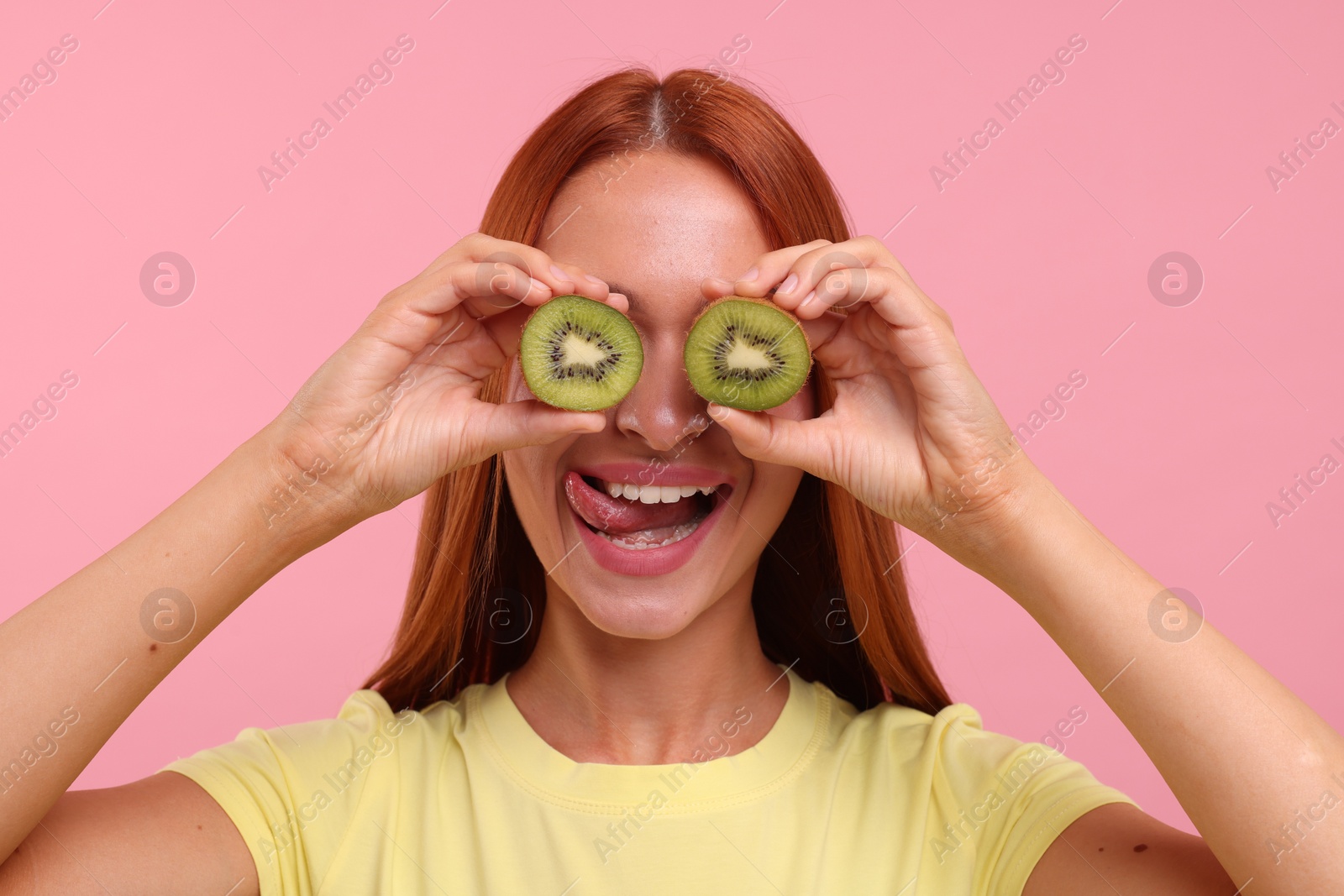 Photo of Funny woman covering eyes with halves of fresh kiwi and showing tongue on pink background