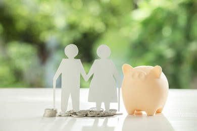 Photo of Pension savings. Figure of senior couple, piggy bank and coins on white table against blurred green background