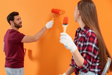 Photo of Man painting orange wall and woman with brush. Interior design