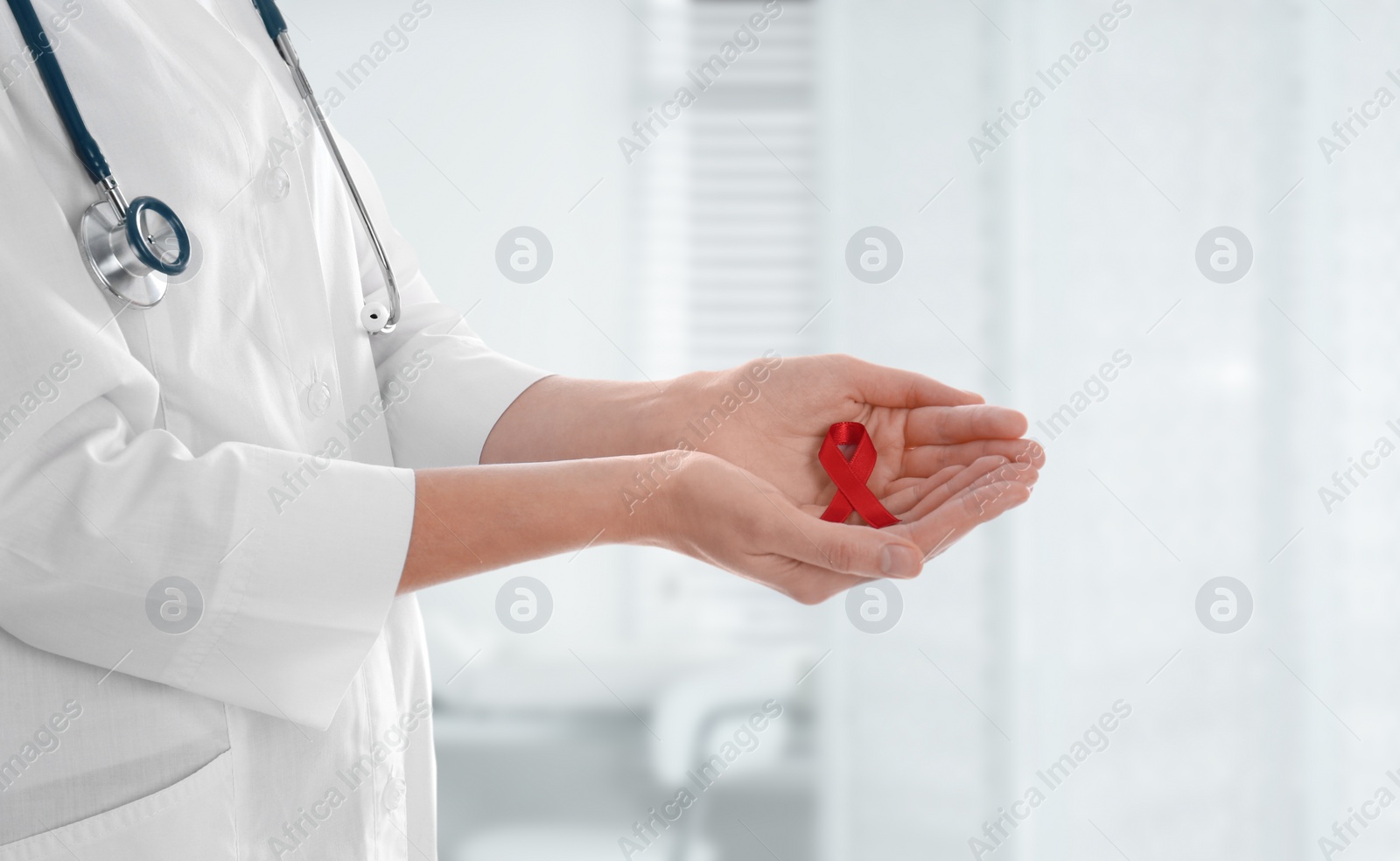 Image of Doctor holding red awareness ribbon on blurred background, closeup. World AIDS disease day