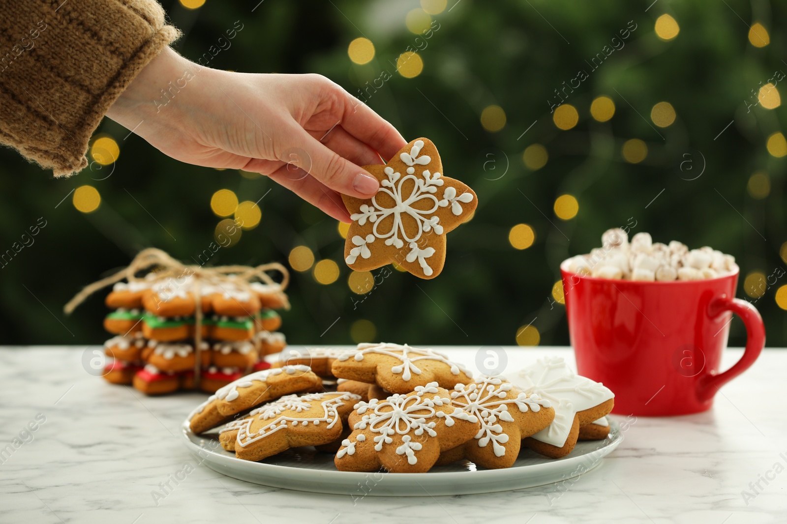 Photo of Woman with decorated cookie at table against blurred Christmas lights, closeup