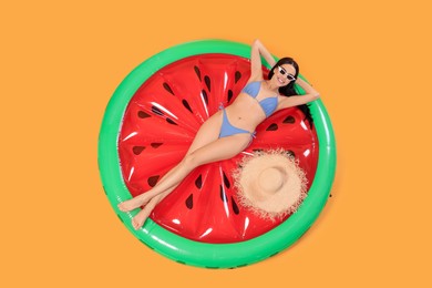 Photo of Happy young woman with beautiful suntan, sunglasses and hat on inflatable mattress against orange background, top view